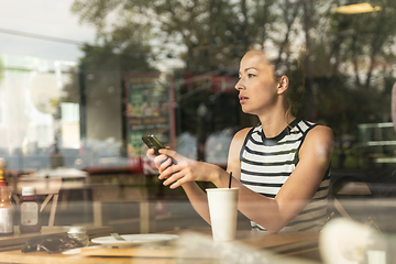 Image showing Thoughtful caucasian woman holding mobile phone while looking through the coffee shop window during coffee break.