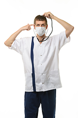 Image showing A masked doctor with a phonendoscope listens to his head, with the other hand he applied an impromptu pistol to his temple, isolated on a white background