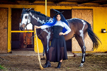 Image showing A girl dressed as a witch stands with a broom near a horse on which a skeleton is drawn