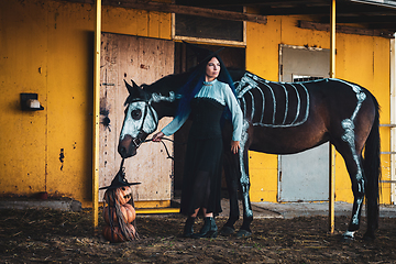 Image showing A girl dressed as a witch on a farm, a horse with a skeleton is drawn next to it, as well as three pumpkins in the shape of an evil man