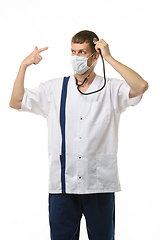 Image showing Masked doctor using a phonendoscope listens to his head, with the other hand making a pistol gesture points to his head, isolated on white background