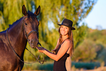 Image showing Happy girl in a hat and a horse in the rays of the warm setting sun looked into the frame