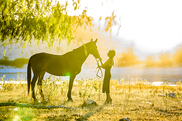 Image showing A girl stands in front of a horse on the shore of a lake and is flooded with warm sunbeams at sunset