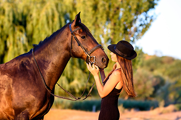 Image showing A girl in a black hat in the rays of the setting sun looks happily at a horse