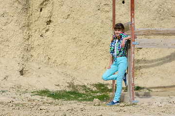 Image showing Stylish cute girl in a plaid shirt and jeans leans on a wooden structure against a background of a rock