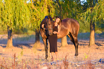 Image showing A girl with a horse stands against the background of trees, the rays of the setting sun fall on them