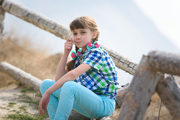 Image showing A girl sits on a fence made of a wooden blockhouse and, with a blade of grass in her mouth, looks into the frame