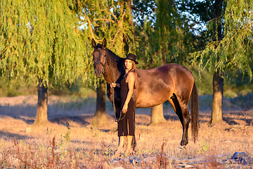 Image showing A slender beautiful girl in a black dress and hat walks with a horse