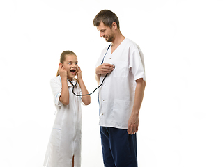 Image showing The doctor put the head of the phonendoscope to his heart, a girl in a medical coat listens with surprise to the doctor\'s heart