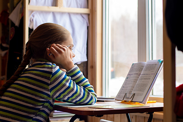 Image showing The schoolgirl carefully reads the task in the textbook while sitting at the table by the window at home