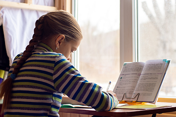 Image showing Schoolgirl sits at a table by the window and does her homework, view from the back