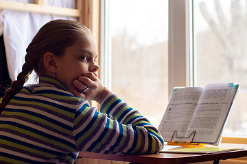 Image showing A schoolgirl sits at a table by the window and does her homework, the girl was distracted in thought and looked towards the frame