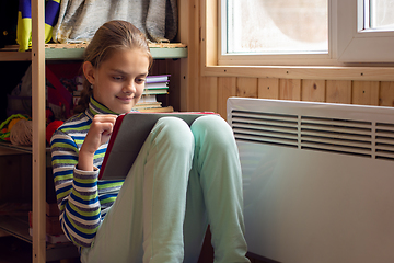 Image showing A girl plays in a tablet sitting next to a radiator by a window in a country house