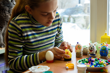 Image showing A girl sits at a table by the window and paints Easter eggs with a brush and paints