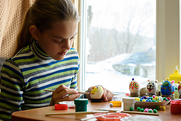 Image showing A girl sits at a table by the window and paints Easter eggs with a brush and paints