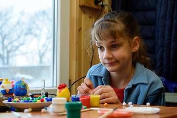 Image showing The girl funny thought painting Easter eggs while sitting at the table by the window