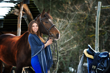 Image showing A beautiful girl in a blue dress and a stole stands next to a horse on the background of a forest and wooden buildings