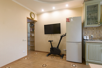 Image showing A fragment of the interior of the living room combined with the kitchen, a TV hangs on the wall, an exercise bike and a refrigerator are located nearby