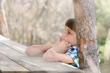 Image showing A ten-year-old girl sits at a wooden table in the forest and thinks