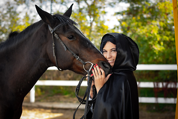 Image showing A girl in a black cloak hugs the muzzle of a horse against the background of trees and a fence