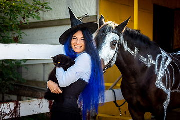 Image showing A girl dressed as a witch holds a black cat in her arms and stands by a corral on a farm next to a horse