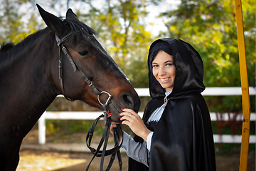 Image showing Happy girl in medieval clothes and a black cloak stands with a horse horse on the background of trees and a fence