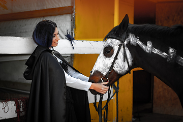 Image showing A beautiful girl in medieval style clothes looks at a horse painted with white paint with a painted skeleton