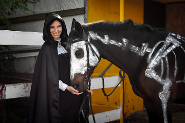 Image showing A beautiful girl in medieval style clothes looks happily into the frame and feeds a horse painted with white paint with a painted skeleton