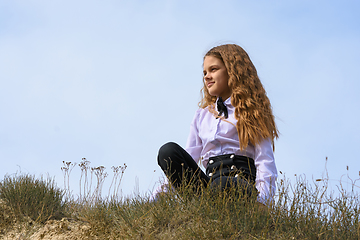 Image showing A girl in a white shirt with a bow tie sits on the ground in a field against the background of the sky