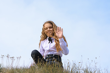 Image showing A girl in a white shirt with a bow tie sits on the ground in a field against the background of the sky and waves a pen to the frame