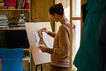 Image showing The artist paints a drawing of a cat with acrylic paints on an easel, in a small workshop by the window