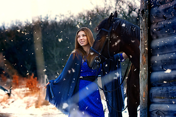 Image showing Beautiful girl in a blue stole stands next to a horse near wooden buildings on a snowy winter day