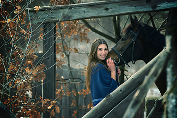 Image showing A beautiful girl in a blue stole stands next to a horse on the background of wooden buildings