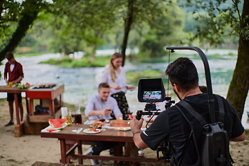 Image showing a professionally equipped cameraman is filming a group of people having dinner by the river.