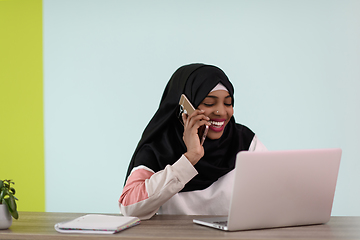 Image showing afro muslim woman wearing a hijab sits smiling in her home office and talking on smartphone