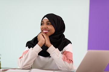 Image showing afro muslim woman wearing a hijab sits smiling in her home office and uses a laptop