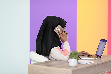 Image showing afro muslim woman wearing a hijab sits smiling in her home office and talking on smartphone