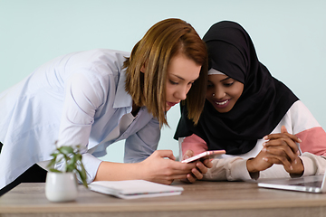 Image showing afro girl with a hijab and a European woman use a cell phone and laptop in their home office