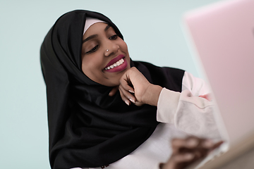 Image showing afro muslim woman wearing a hijab sits smiling in her home office and uses a laptop