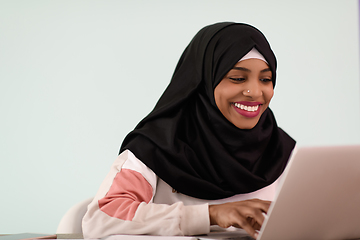 Image showing afro muslim woman wearing a hijab sits smiling in her home office and uses a laptop