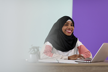 Image showing afro muslim woman wearing a hijab sits smiling in her home office and uses a laptop