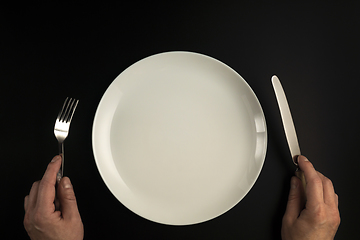 Image showing Hands with fork and knife and white plate on black background in flat lay