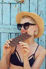 Image showing Young attractive woman in sunglasses and in straw hat eating chocolate on a blue background. Film efect