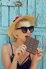 Image showing Young attractive woman in sunglasses and in straw hat eating chocolate on a blue background. Film efect