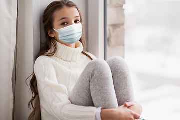 Image showing sad girl in medical mask sitting on sill at home