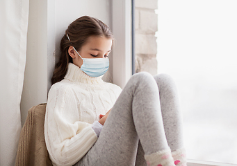 Image showing sad girl in medical mask sitting on sill at home