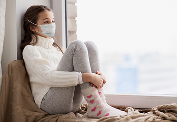 Image showing sad girl in medical mask sitting on sill at home