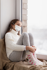 Image showing sad girl in medical mask sitting on sill at home