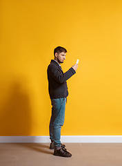 Image showing Young caucasian man using smartphone. Full body length portrait isolated over yellow background.