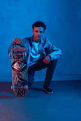 Image showing Caucasian young skateboarder posing on dark neon lighted background
